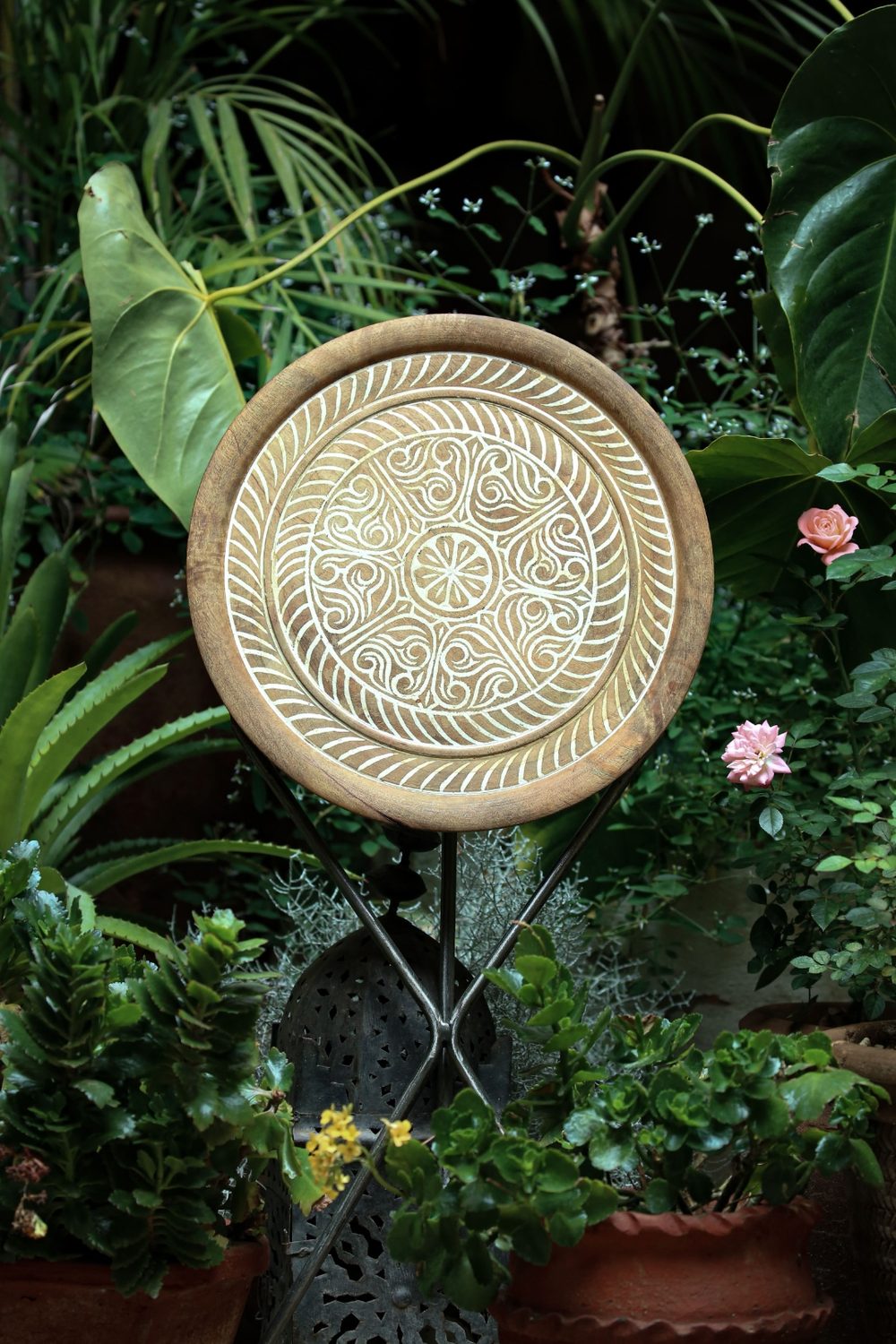 A round hand-carved wooden tabletop on a tripod stand, set among potted plants and roses