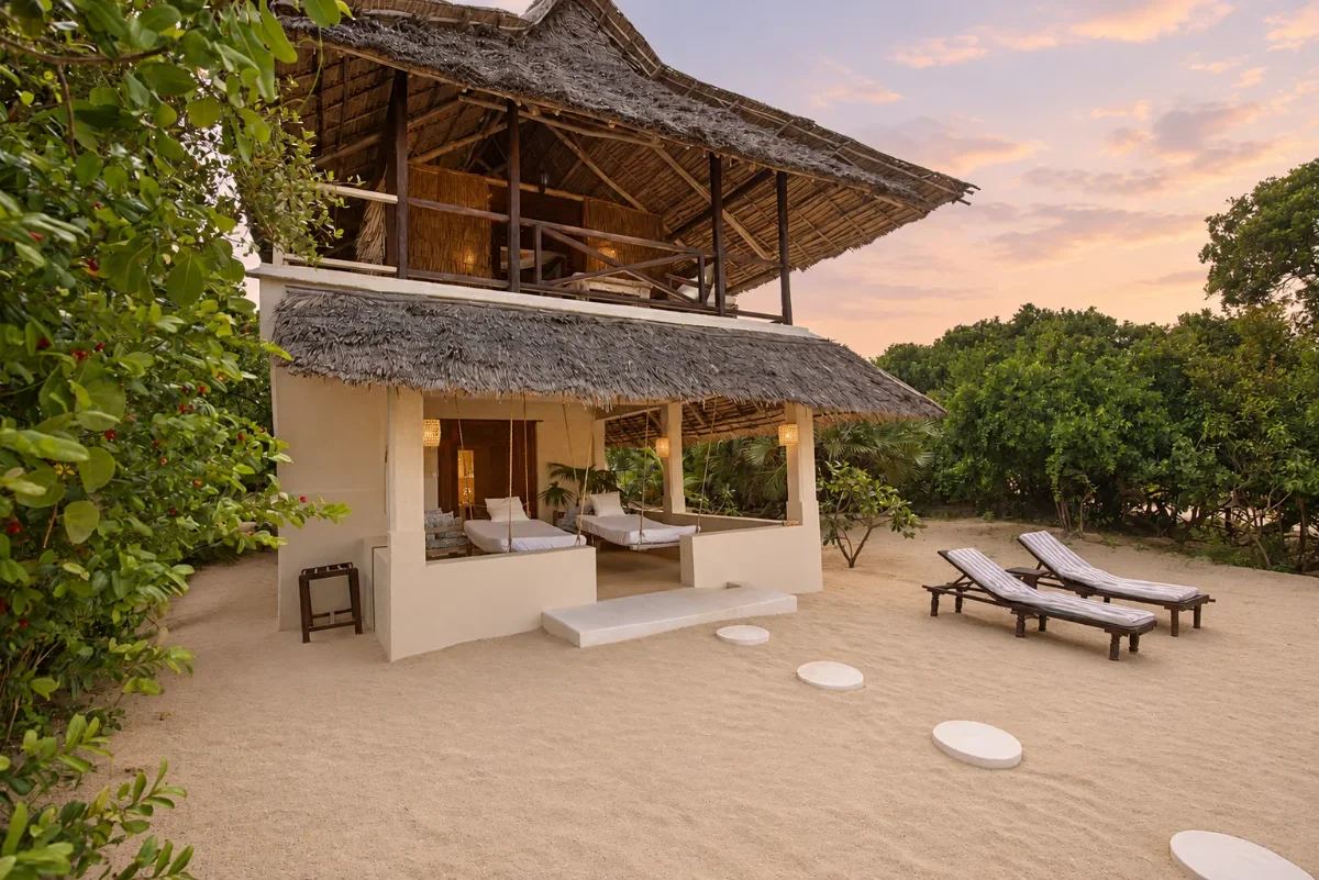 Makai cabana at dusk, two thatched storeys above the sand with loungers and stepping stones