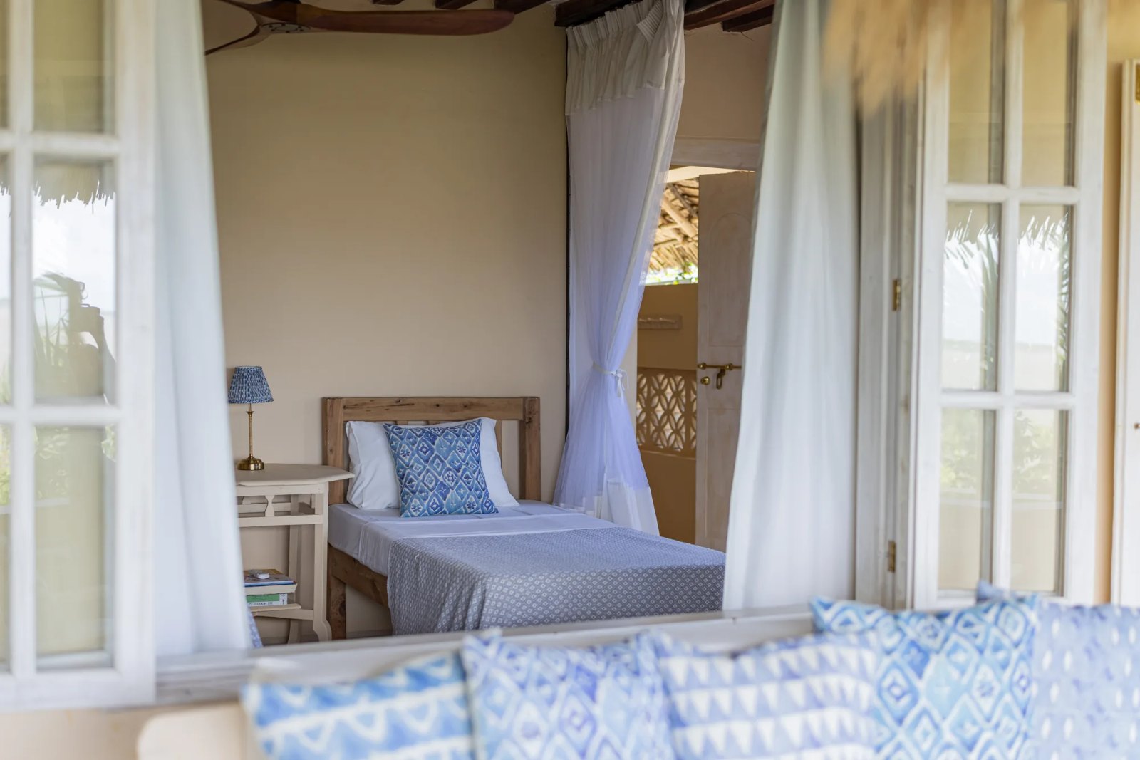 Moana cabana · twin bed glimpsed through the shutters, Bali-blue cushions in the foreground