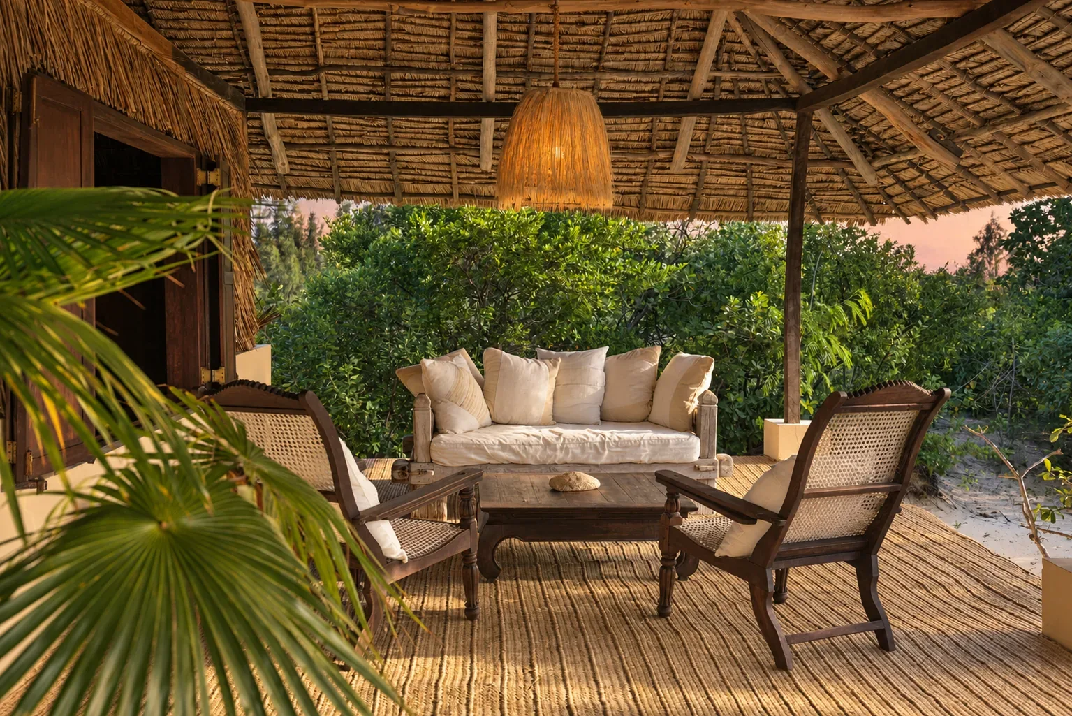 ʻOhana cabana, the verandah lounge under makuti thatch, a daybed and planter chairs at golden hour