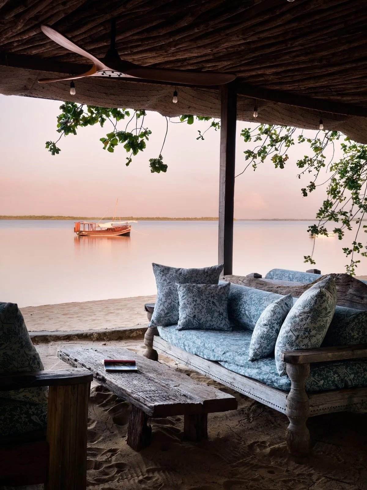 A shaded beach lounge with blue cushions looking out to a dhow at anchor
